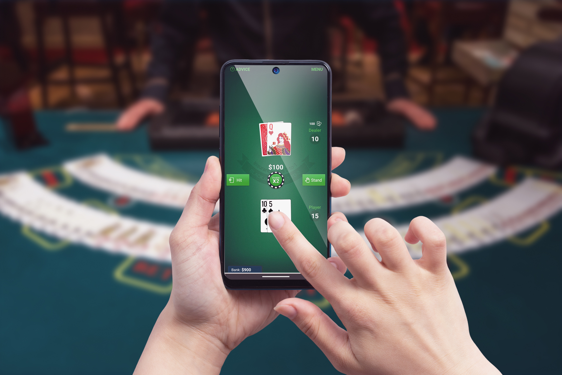 Close-up of a person's hands playing a mobile blackjack game on a smartphone, with a green digital table displaying the player's cards (10 of spades and 5 of clubs) and the dealer's card (Queen of hearts). The player is deciding whether to hit or stand. The background is blurred, suggesting a real casino setting, which emphasises the contrast between digital and traditional gaming environments.